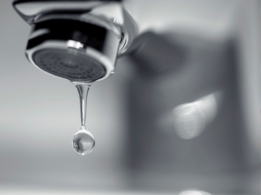 A water droplet falling from a chrome faucet aerator against a blurred gray background