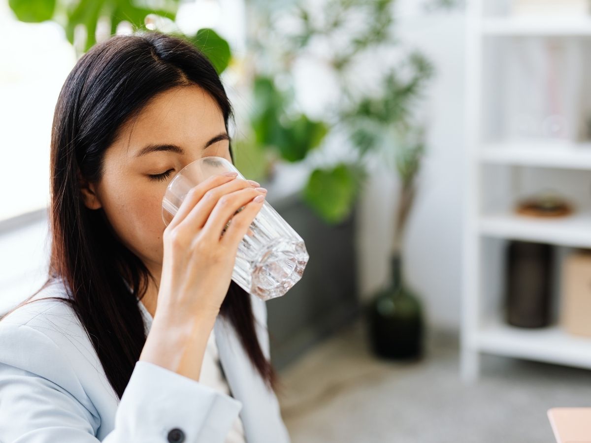 Woman in white blazer drinking water from a glass in a bright office with potted plants