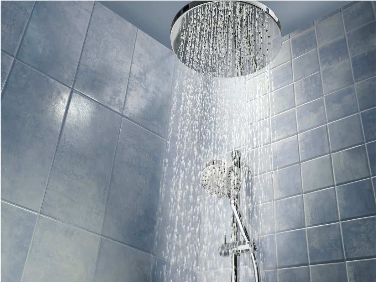 Modern rainfall shower head mounted on ceiling with handheld showerhead below, water cascading against blue-gray tile walls in contemporary bathroom