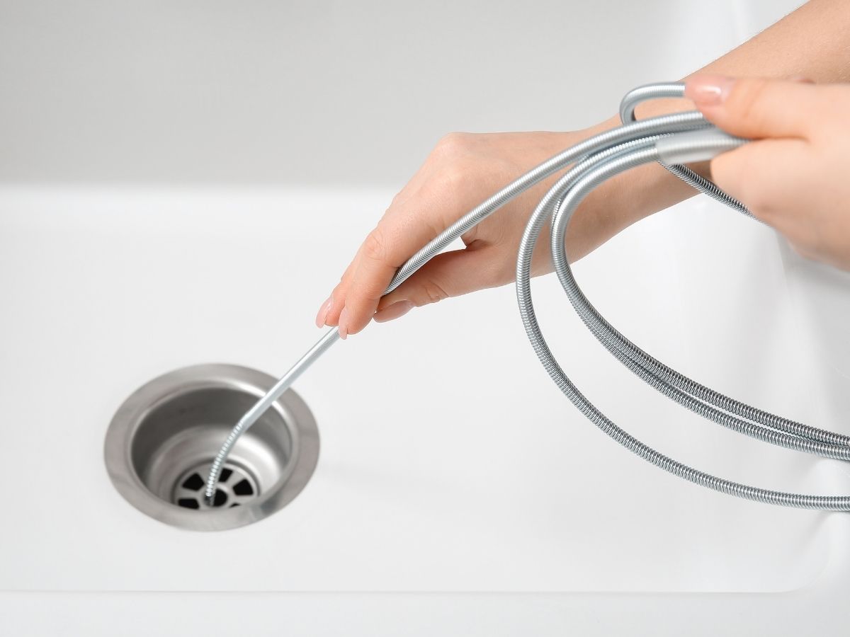 Hand holding a metal drain snake cable being inserted into a bathroom sink drain opening