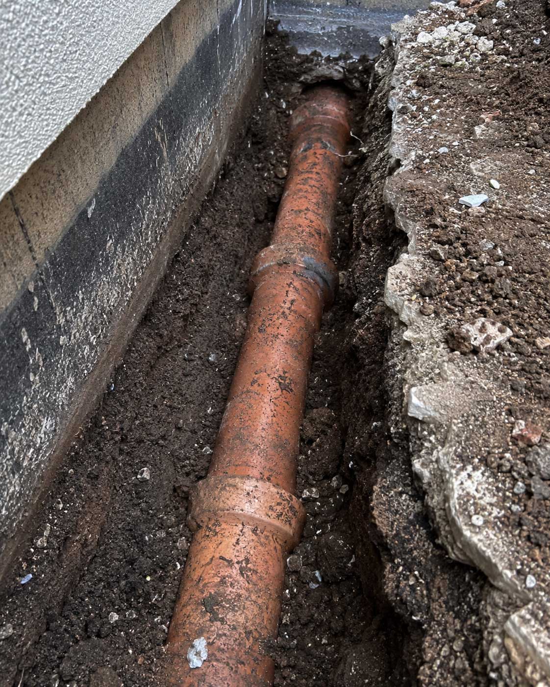 Rusty metal pipe buried in soil between concrete foundation and stone wall in narrow trench