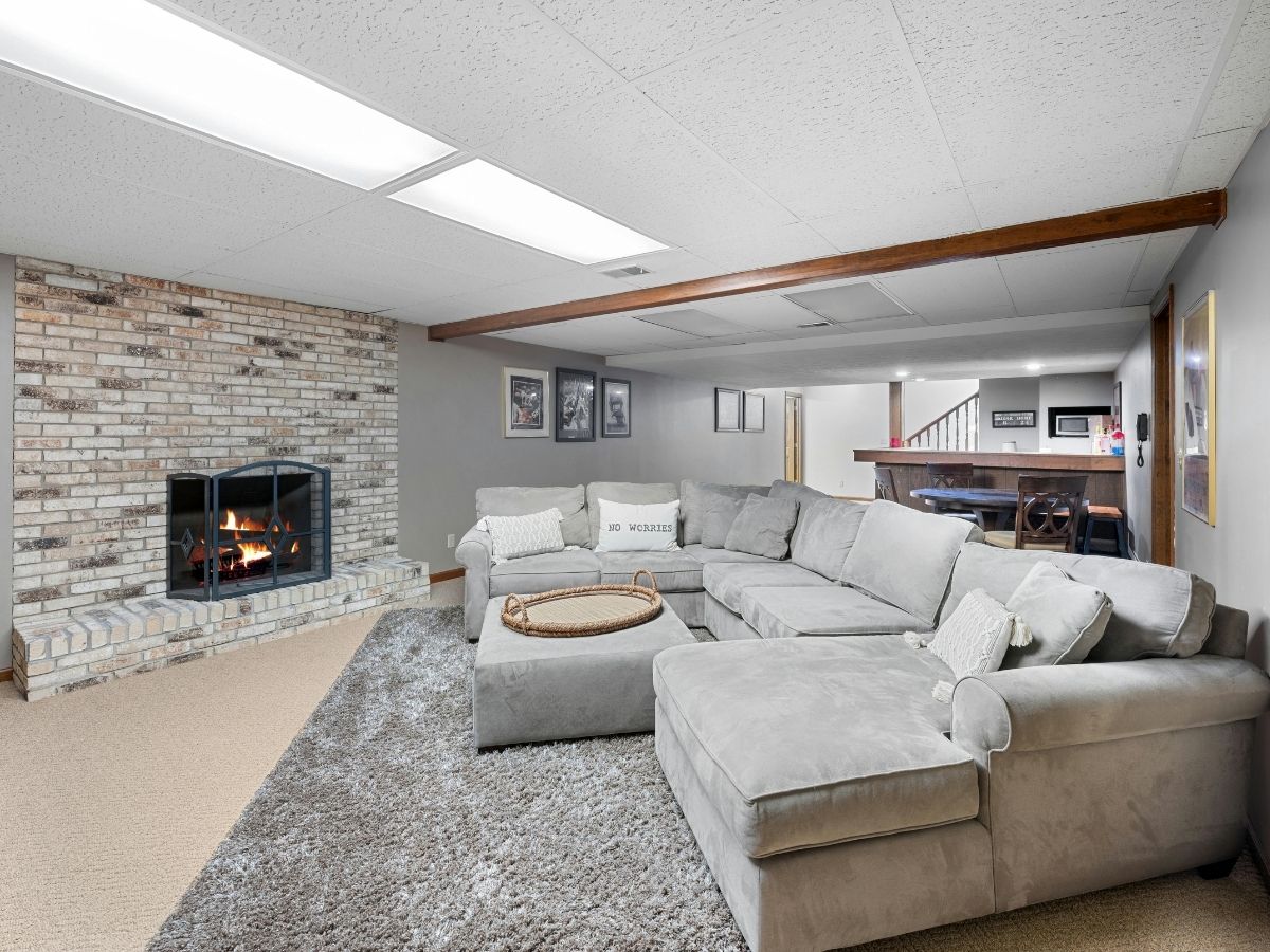 Finished basement with large sectional sofa, fireplace with brick surround, wooden beam accent, and open layout leading to bar area