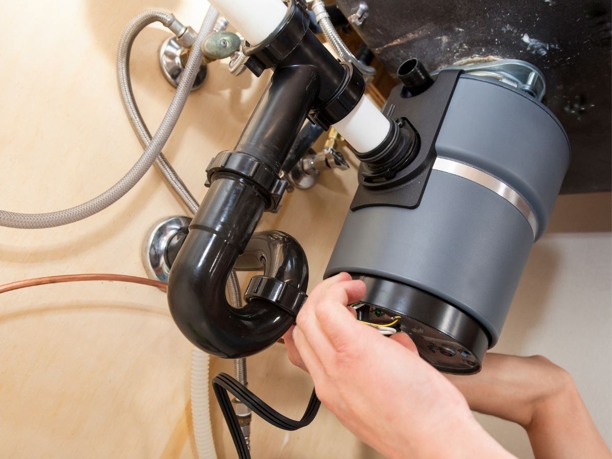 Hand holding a black garbage disposal unit connected to plumbing pipes and hoses under a sink
