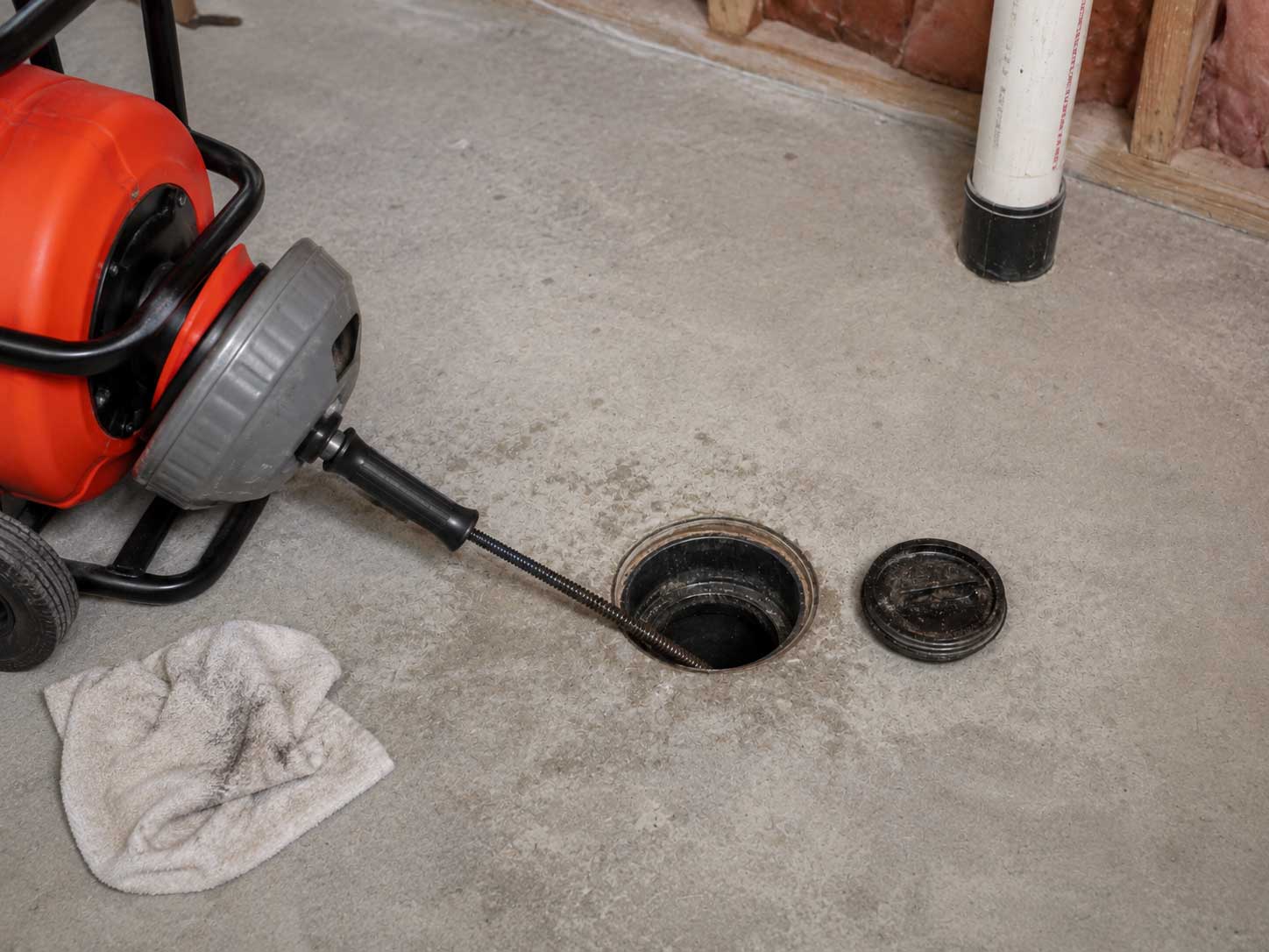Red motorized drain cleaning machine with flexible snake hose positioned over open floor drain, alongside removed drain cover and cloth