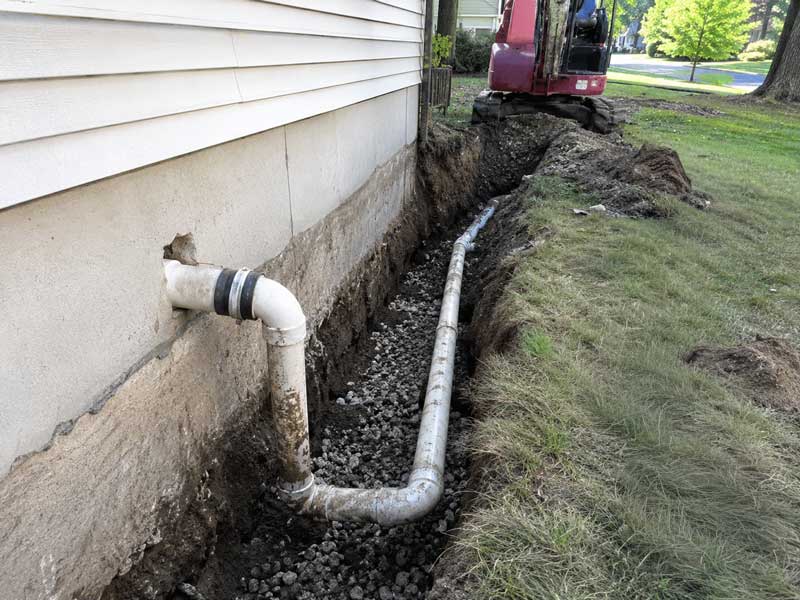 Excavated trench along house foundation showing drainage pipe and gravel base during sewer line installation or repair work