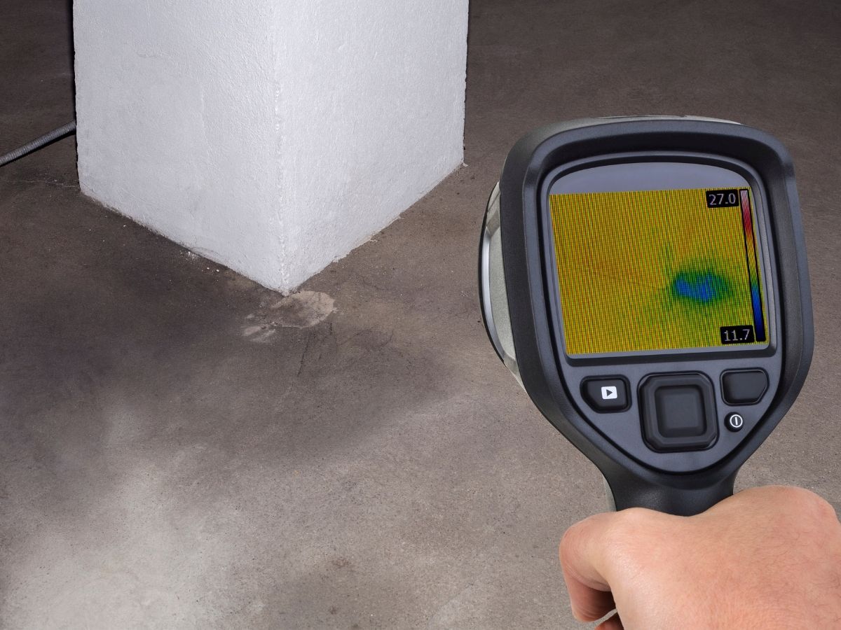 Thermal imaging camera detecting moisture leak under white foam insulation on concrete floor, displaying temperature differential in yellow and blue heat signature