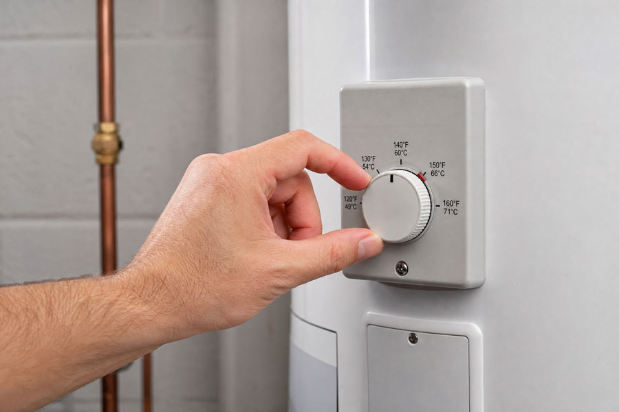 Man's hand adjusting a wall-mounted thermostat dial showing temperature settings in Fahrenheit and Celsius, with copper heating pipes visible in background