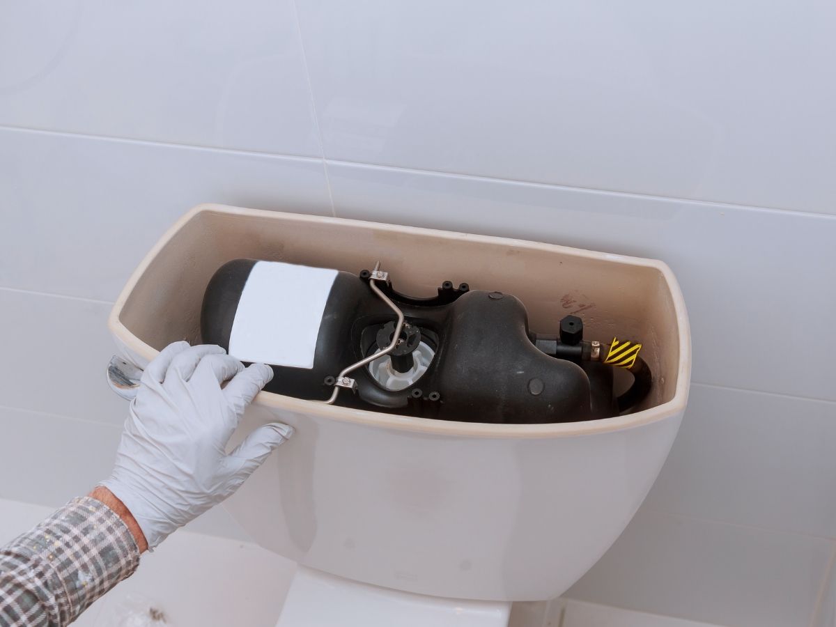 Gloved hand installing white fill valve and black flush mechanism inside open toilet tank