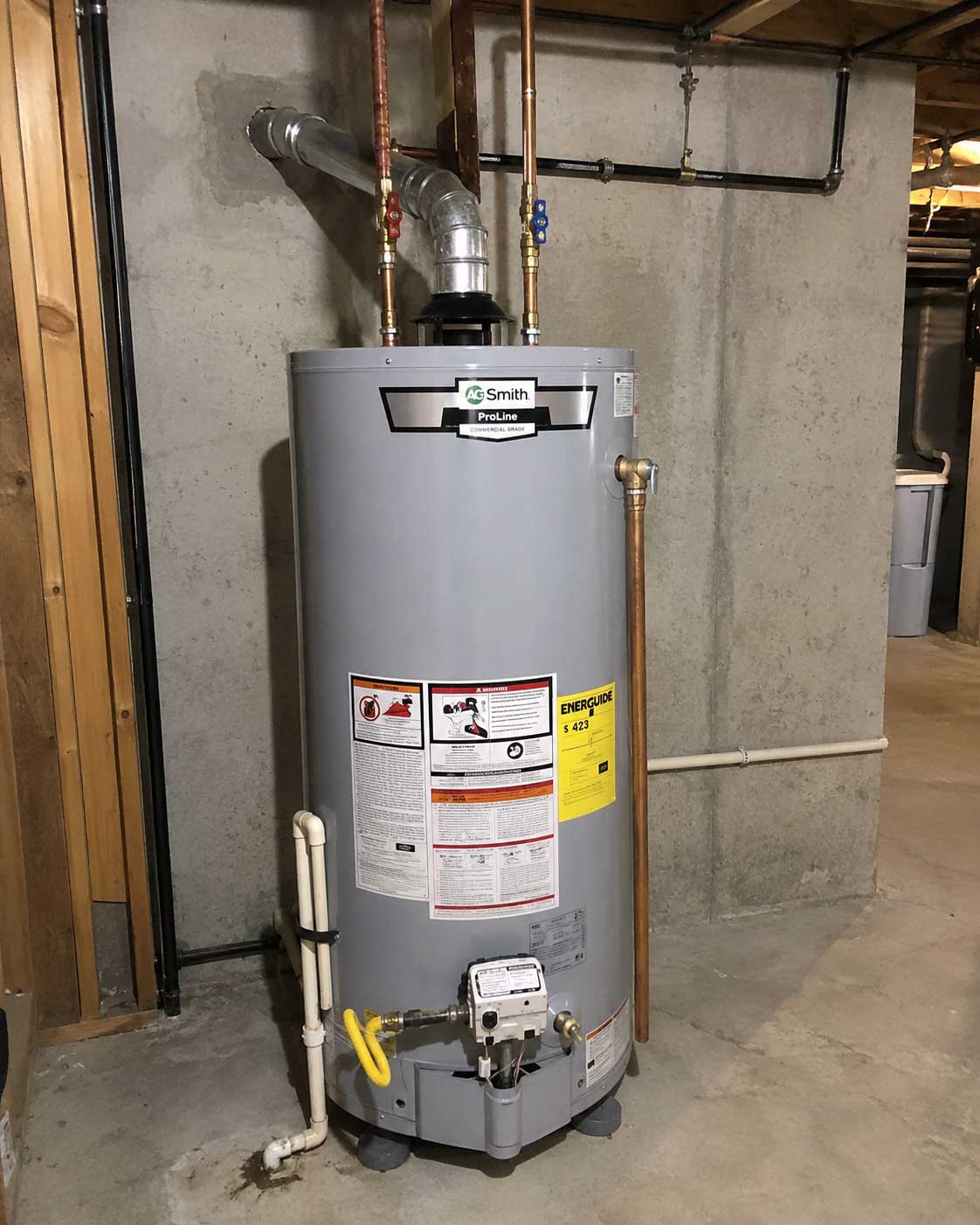 A.O. Smith ProLine gas water heater installed in a basement, with copper and black pipes connected to the top, yellow valve at the bottom, and an EnergyGuide label on the front