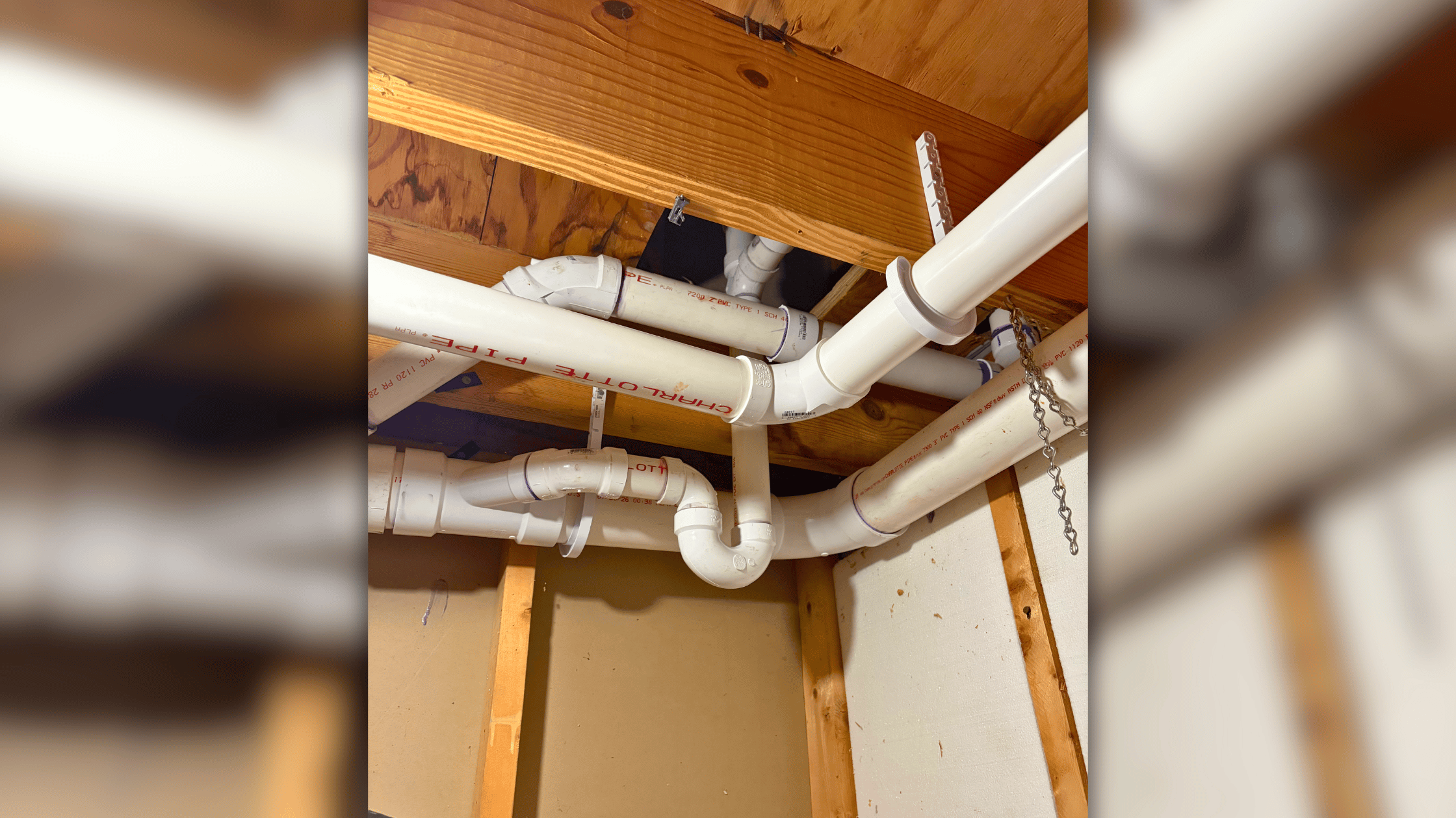 White PVC pipes and plumbing fixtures mounted under wooden ceiling beams and joists in an unfinished interior space