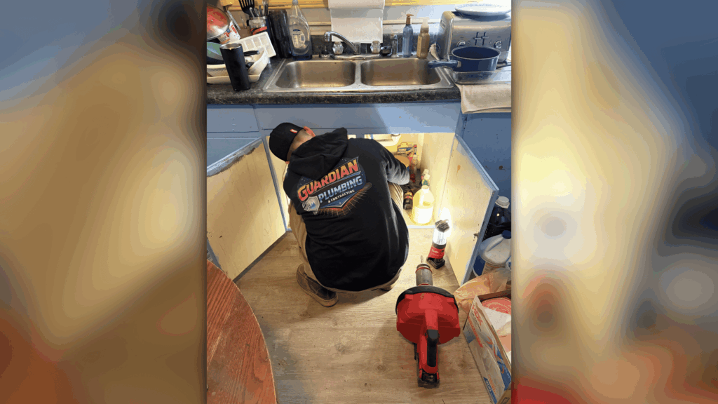Plumber in Guardian Plumbing shirt working under kitchen sink cabinet with tools and supplies visible on floor