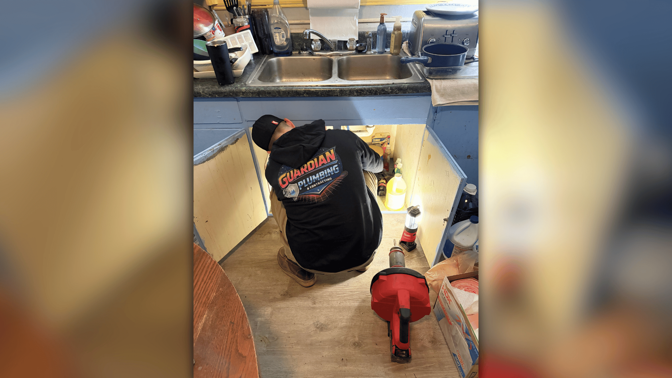 Plumber in Guardian Plumbing shirt working under kitchen sink cabinet with tools and supplies visible on floor