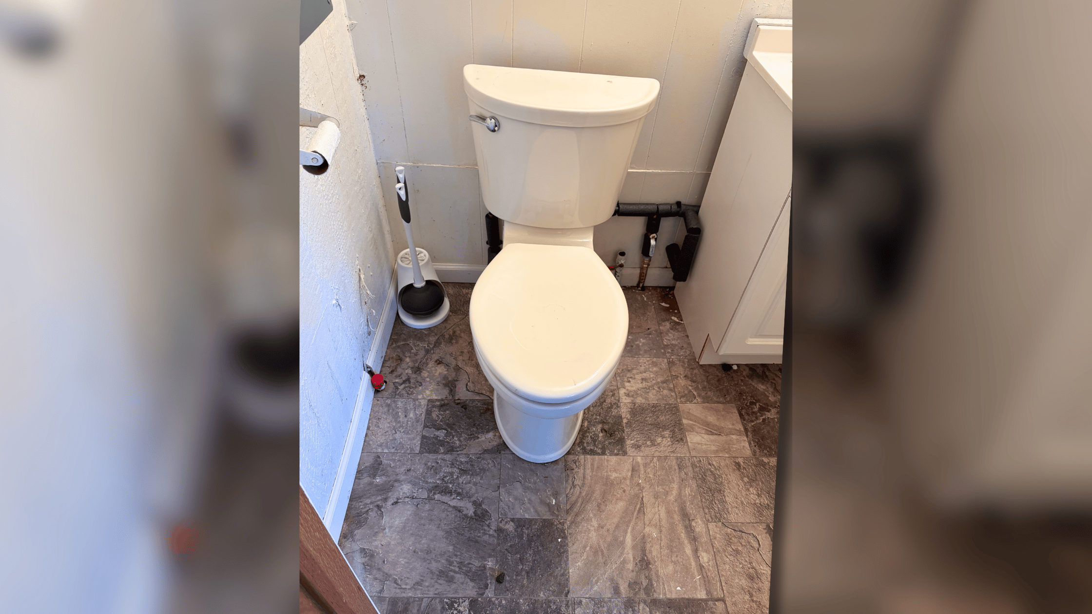 A clean white toilet with seat down in a small bathroom with gray tile flooring and white walls, featuring a plunger on the left side
